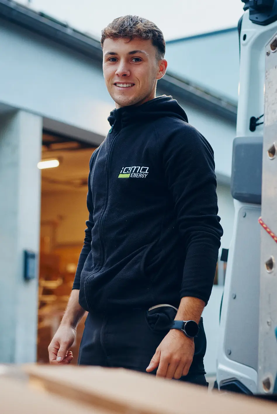 Lächelnder Mitarbeiter im iQma energy Pullover am Transporter vor der Lagerhalle